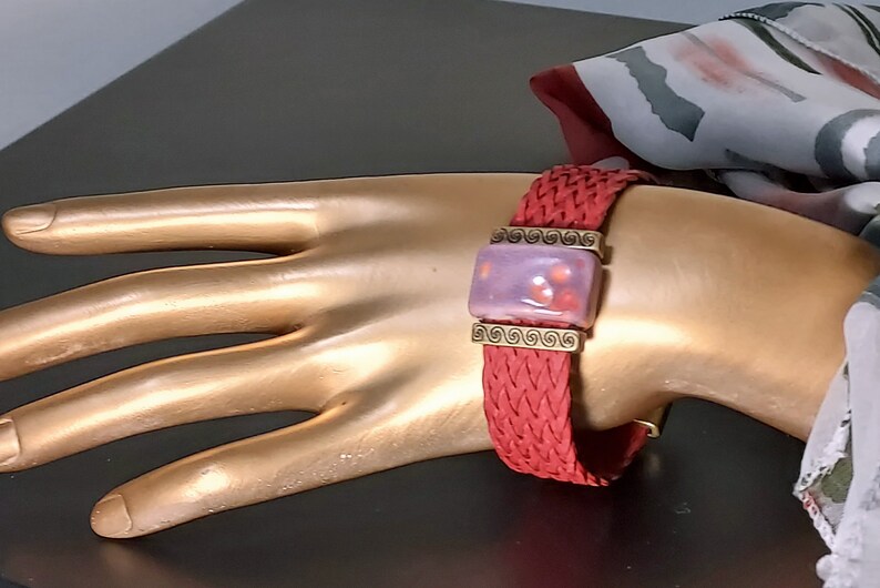 May include: A red braided bracelet with a rectangular lavender and red centerpiece. The centerpiece is framed with gold-colored metal and features a swirl design. The bracelet is displayed on a gold-colored hand.