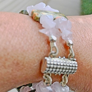 May include: A close-up of a bracelet featuring a mix of materials. The bracelet has a silver clasp and is adorned with light pink, irregular-shaped stones and a larger, patterned stone. The design incorporates a floral element with white, petal-like accents.