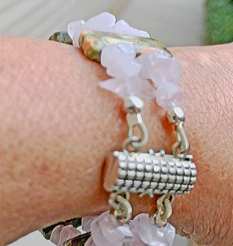 May include: A close-up of a bracelet featuring a mix of materials. The bracelet has a silver clasp and is adorned with light pink, irregular-shaped stones and a larger, patterned stone. The design incorporates a floral element with white, petal-like accents.