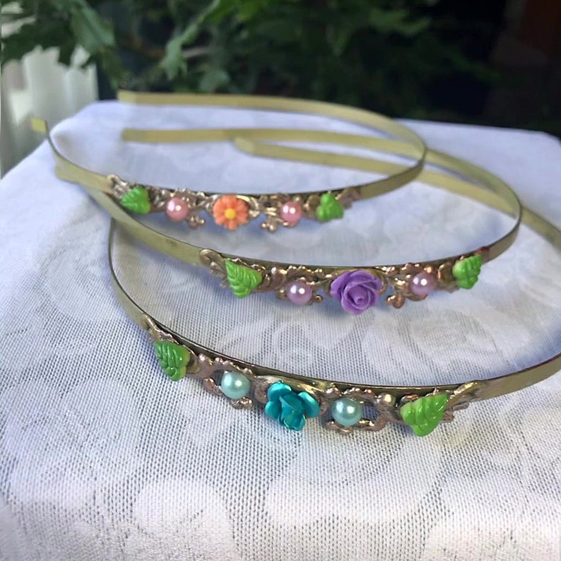 May include: Two gold metal headbands with green leaves, pink and white pearls, and colorful flowers. The flowers are orange, purple, and blue.