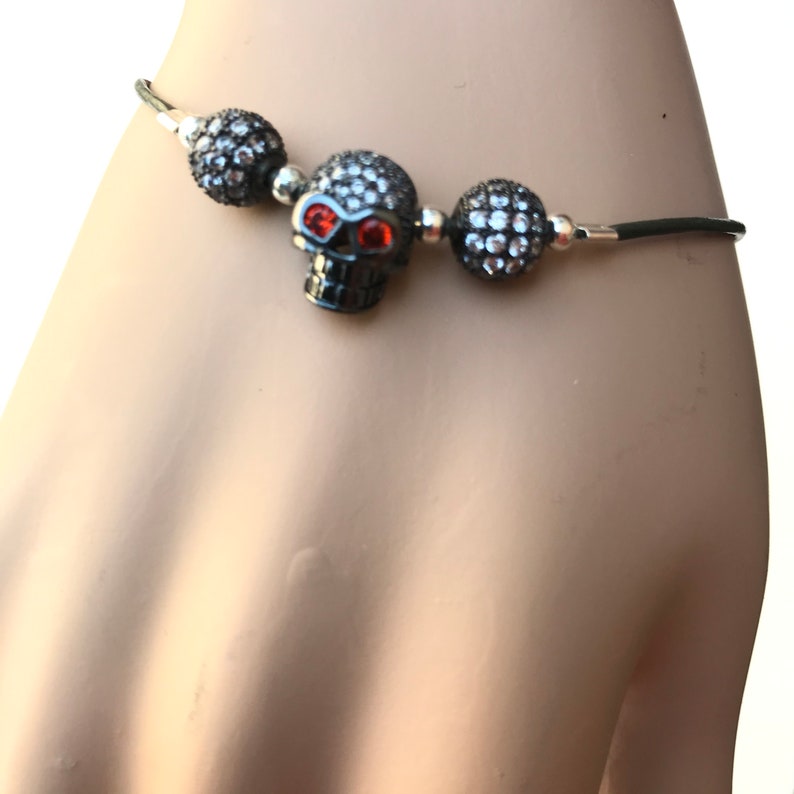 Op de afbeelding: Zwarte schedelarmband met rode oogaccenten en met kristal bedekte kralen. De schedel heeft een donkere afwerking en is gecentreerd tussen twee met kristal bedekte kralen. De armband is geregen op een zwart koord.