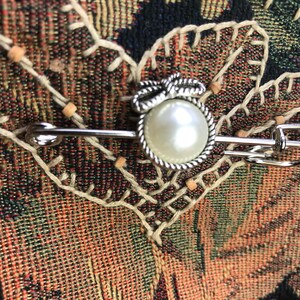 May include: A silver-toned brooch with a large, round, faux pearl center. The pearl is surrounded by a twisted rope-like border and a bow detail. The brooch is set against a textured, multi-colored fabric background.