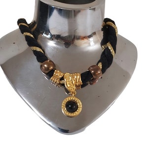 May include: A black and gold braided necklace with a gold pendant featuring a black stone. The necklace has gold accents and bronze-colored beads. The pendant is round with a gold frame.