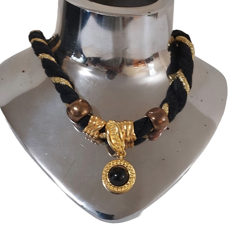 May include: A black and gold braided necklace with a gold pendant featuring a black stone. The necklace has gold accents and bronze-colored beads. The pendant is round with a gold frame.