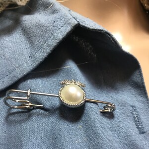 May include: A silver-toned safety pin with a faux pearl pendant. The pendant is a round, off-white pearl surrounded by a decorative silver frame. The pin is fastened on a light blue fabric.