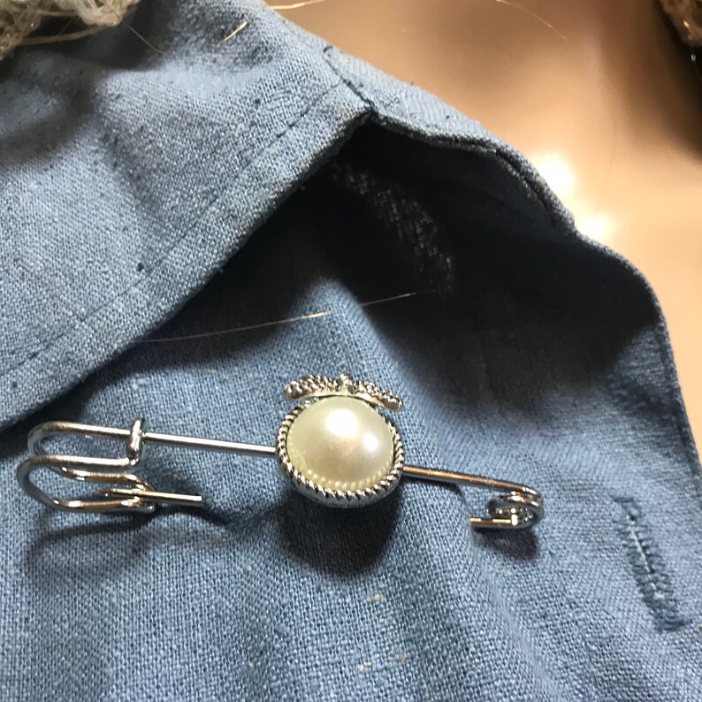 May include: A silver-toned safety pin with a faux pearl pendant. The pendant is a round, off-white pearl surrounded by a decorative silver frame. The pin is fastened on a light blue fabric.