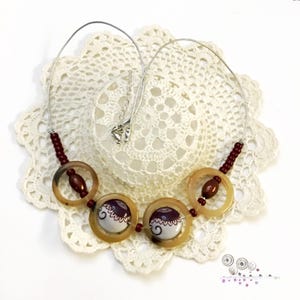 May include: A necklace featuring a silver-toned wire cord with dark red beads and four translucent tan rings. Two of the rings contain white circular inserts decorated with dark purple patterns and scalloped edges, while the others hold dark red beads.