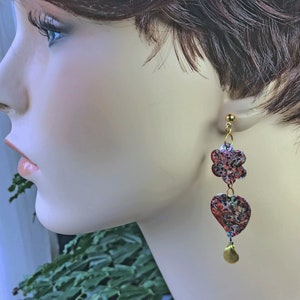 May include: Gold-toned dangle earrings featuring a floral and heart design. The earrings have a textured, colorful pattern with red, blue, and green hues. A small gold teardrop hangs at the bottom of each earring.
