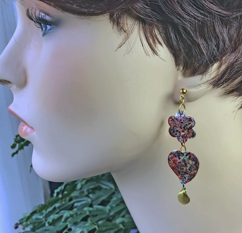May include: Gold-toned dangle earrings featuring a floral and heart design. The earrings have a textured, colorful pattern with red, blue, and green hues. A small gold teardrop hangs at the bottom of each earring.