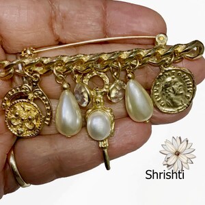 May include: A gold-toned brooch featuring a chain design with dangling charms. The charms include faux pearls, clear crystals, and a coin-like pendant. The brooch is held in a hand, with the brand name "Shrishti" visible.