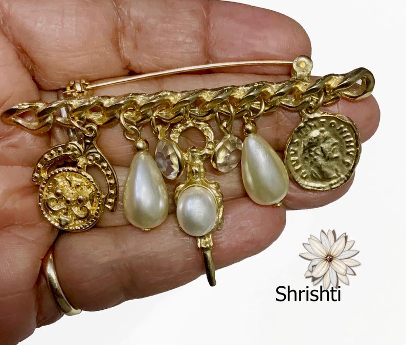 May include: A gold-toned brooch featuring a chain design with dangling charms. The charms include faux pearls, clear crystals, and a coin-like pendant. The brooch is held in a hand, with the brand name "Shrishti" visible.