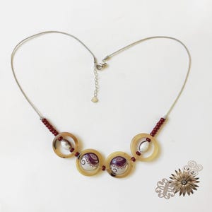 May include: A necklace featuring a thin silver-tone wire chain with a lobster clasp and extender. The centerpiece consists of four circular tan resin rings, two of which contain white and purple patterned circular inserts, accented by small red beads.