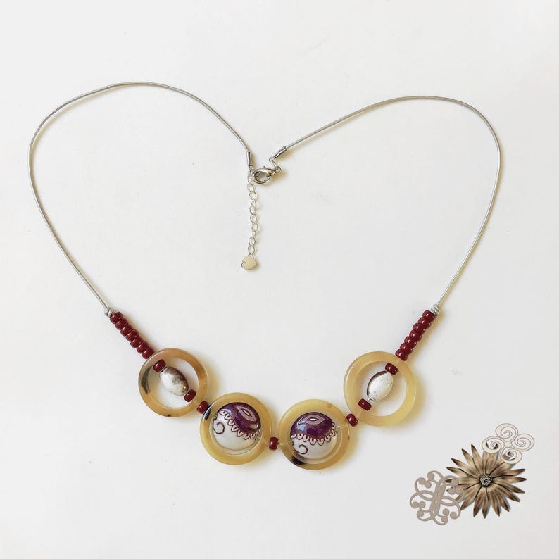 May include: A necklace featuring a thin silver-tone wire chain with a lobster clasp and extender. The centerpiece consists of four circular tan resin rings, two of which contain white and purple patterned circular inserts, accented by small red beads.
