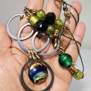 May include: A handcrafted necklace featuring a mix of materials and colors. The necklace includes silver-toned metal rings, green and black beads, and a pendant with a blue and gold glass orb. The necklace is held in a hand.