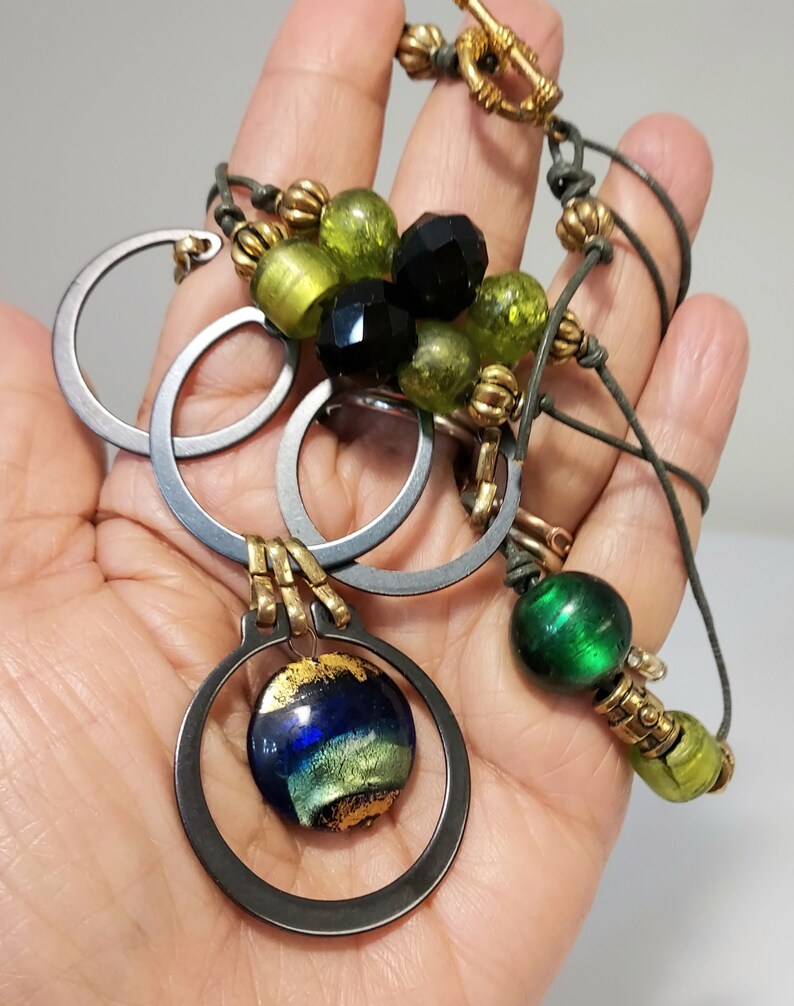 May include: A handcrafted necklace featuring a mix of materials and colors. The necklace includes silver-toned metal rings, green and black beads, and a pendant with a blue and gold glass orb. The necklace is held in a hand.