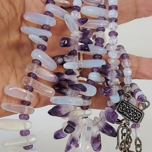 May include: A multi-strand necklace featuring a combination of amethyst and opalescent beads. The necklace has a silver-toned clasp and chain. The beads are in shades of purple, lavender, and white. The necklace is a piece of jewelry.