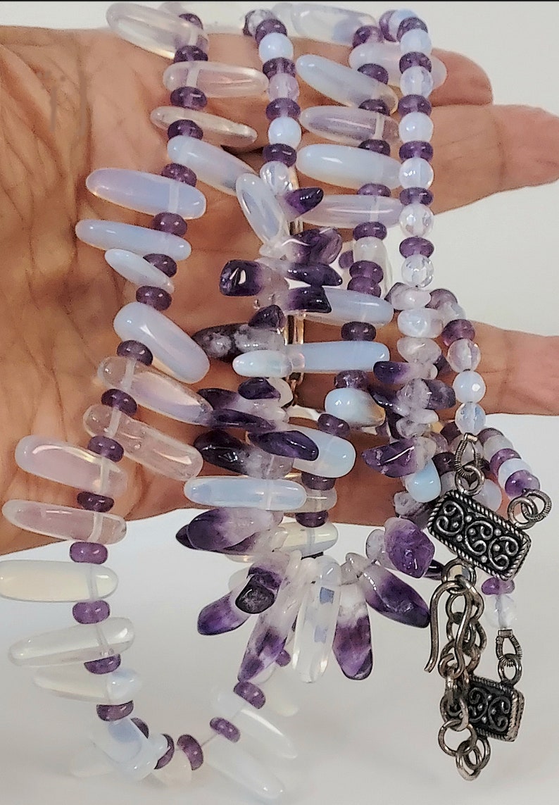 May include: A multi-strand necklace featuring a combination of amethyst and opalescent beads. The necklace has a silver-toned clasp and chain. The beads are in shades of purple, lavender, and white. The necklace is a piece of jewelry.