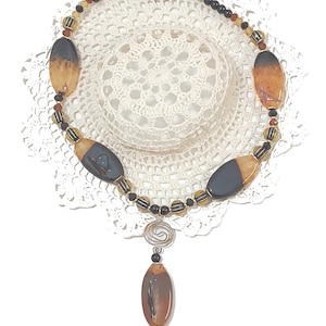 May include: A handcrafted necklace featuring large, oval-shaped beads in shades of brown and black, interspersed with smaller, round beads. The necklace has a silver-tone clasp and a decorative pendant with a spiral design and a dangling bead cluster. The necklace is displayed on a white lace doily.