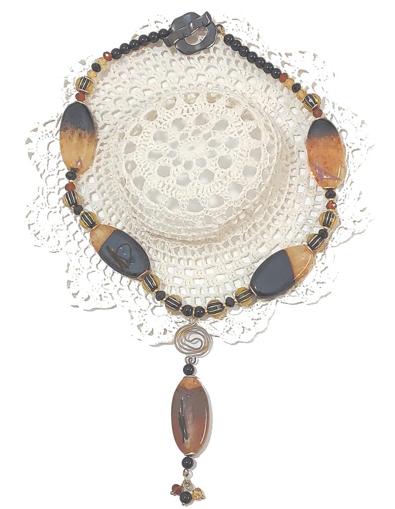 May include: A handcrafted necklace featuring large, oval-shaped beads in shades of brown and black, interspersed with smaller, round beads. The necklace has a silver-tone clasp and a decorative pendant with a spiral design and a dangling bead cluster. The necklace is displayed on a white lace doily.