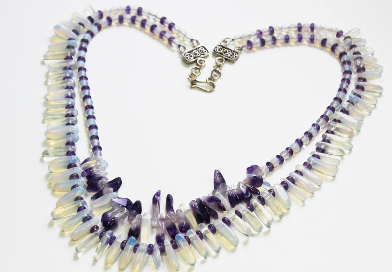 May include: A layered necklace featuring a heart-shaped design. The necklace combines white, teardrop-shaped beads with purple, rounded beads. The necklace is secured with a silver clasp. The necklace is a piece of jewelry.