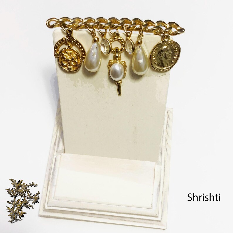 May include: A gold-toned charm bracelet with various pendants, including a floral design, teardrop pearls, and a coin. The charms hang from a chain. The background is a white display stand.