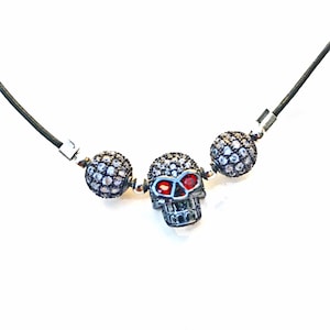 Op de afbeelding: Zwarte koordketting met een zilveren schedel hanger met rode oogaccenten, geflankeerd door twee zilverkleurige, met kristallen bezette kogelkralen. De ketting heeft zilveren sluitingen en staat tegen een witte achtergrond.