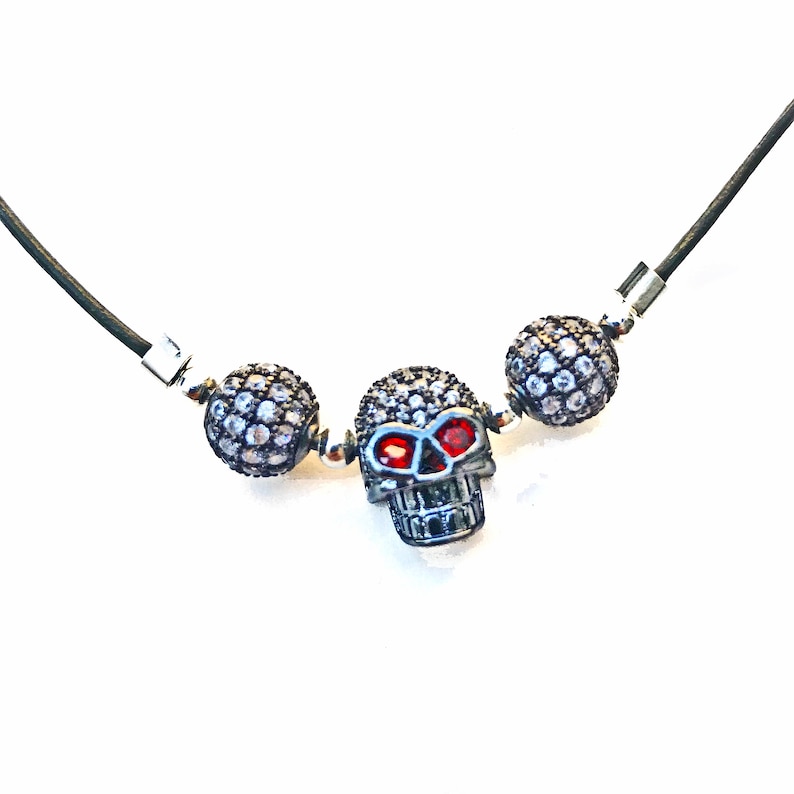 Op de afbeelding: Zwarte koordketting met een zilveren schedel hanger met rode oogaccenten, geflankeerd door twee zilverkleurige, met kristallen bezette kogelkralen. De ketting heeft zilveren sluitingen en staat tegen een witte achtergrond.