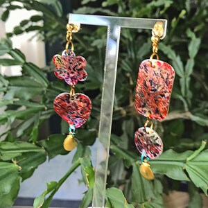 May include: Dangle earrings with a floral, heart, rectangular, and circular design. The earrings feature a gold-colored chain and accents, with colorful abstract patterns in red, pink, and black. The earrings are displayed on a clear stand.