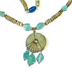 May include: A handcrafted necklace featuring a light green jade pendant with a central hole, wire-wrapped with gold-colored metal. The necklace includes light green and blue-green beads, with a blue oval bead near the clasp. The design is set against a white background.