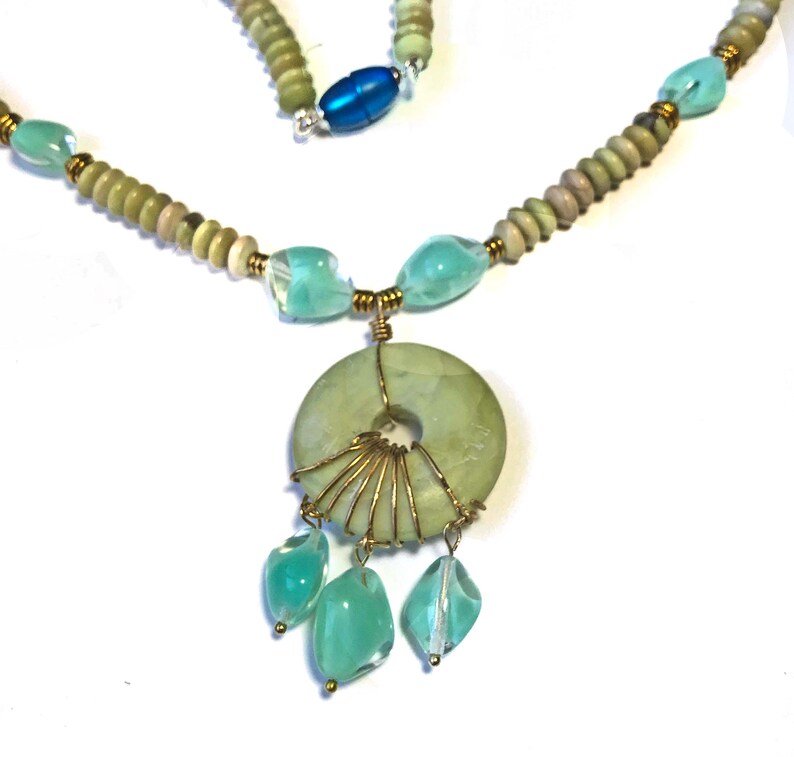 May include: A handcrafted necklace featuring a light green jade pendant with a central hole, wire-wrapped with gold-colored metal. The necklace includes light green and blue-green beads, with a blue oval bead near the clasp. The design is set against a white background.