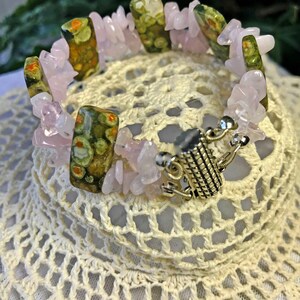May include: A gemstone bracelet featuring a mix of pink rose quartz chips and rectangular green and brown jasper stones. The bracelet has a silver-colored clasp and is displayed on a white, crocheted surface.