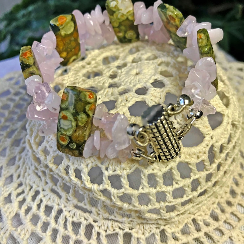 May include: A gemstone bracelet featuring a mix of pink rose quartz chips and rectangular green and brown jasper stones. The bracelet has a silver-colored clasp and is displayed on a white, crocheted surface.