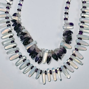 May include: Two layered necklaces. The top layer features a strand of small, clear beads interspersed with purple beads. The bottom layer has teardrop-shaped, iridescent beads with purple accents. The center layer has a mix of purple and clear stones.
