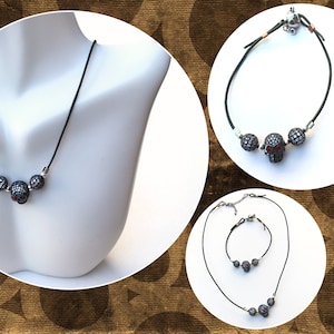 Op de afbeelding: Een zwarte koordketting met drie schedelhangers. De middelste schedel heeft rode ogen. De afbeelding toont ook een bijpassende armband en een ketting- en armbandset, allemaal met hetzelfde schedelontwerp.