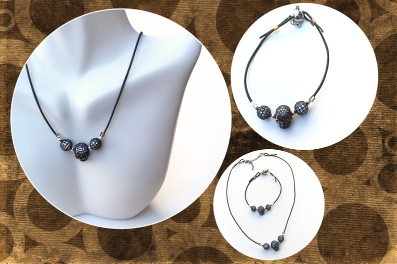 Op de afbeelding: Een zwarte koordketting met drie schedelhangers. De middelste schedel heeft rode ogen. De afbeelding toont ook een bijpassende armband en een ketting- en armbandset, allemaal met hetzelfde schedelontwerp.