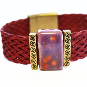 May include: A braided red leather bracelet with a rectangular purple and orange stone centerpiece. The stone is flanked by gold-colored metal accents with a spiral design. The bracelet has a gold-colored clasp.