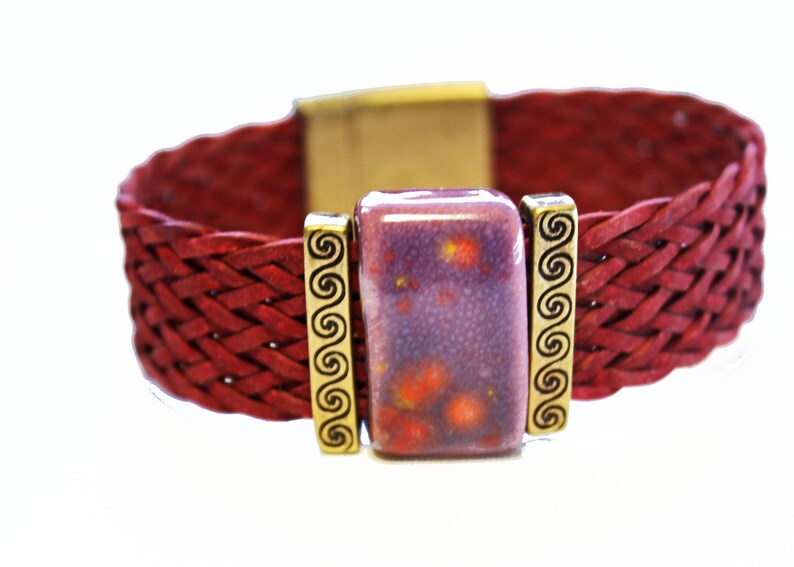 May include: A braided red leather bracelet with a rectangular purple and orange stone centerpiece. The stone is flanked by gold-colored metal accents with a spiral design. The bracelet has a gold-colored clasp.