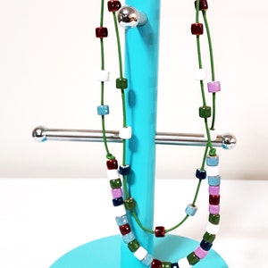 May include: A colorful necklace with square beads in various colors, including red, white, green, and blue, strung on a green cord. The necklace is displayed on a turquoise jewelry stand.