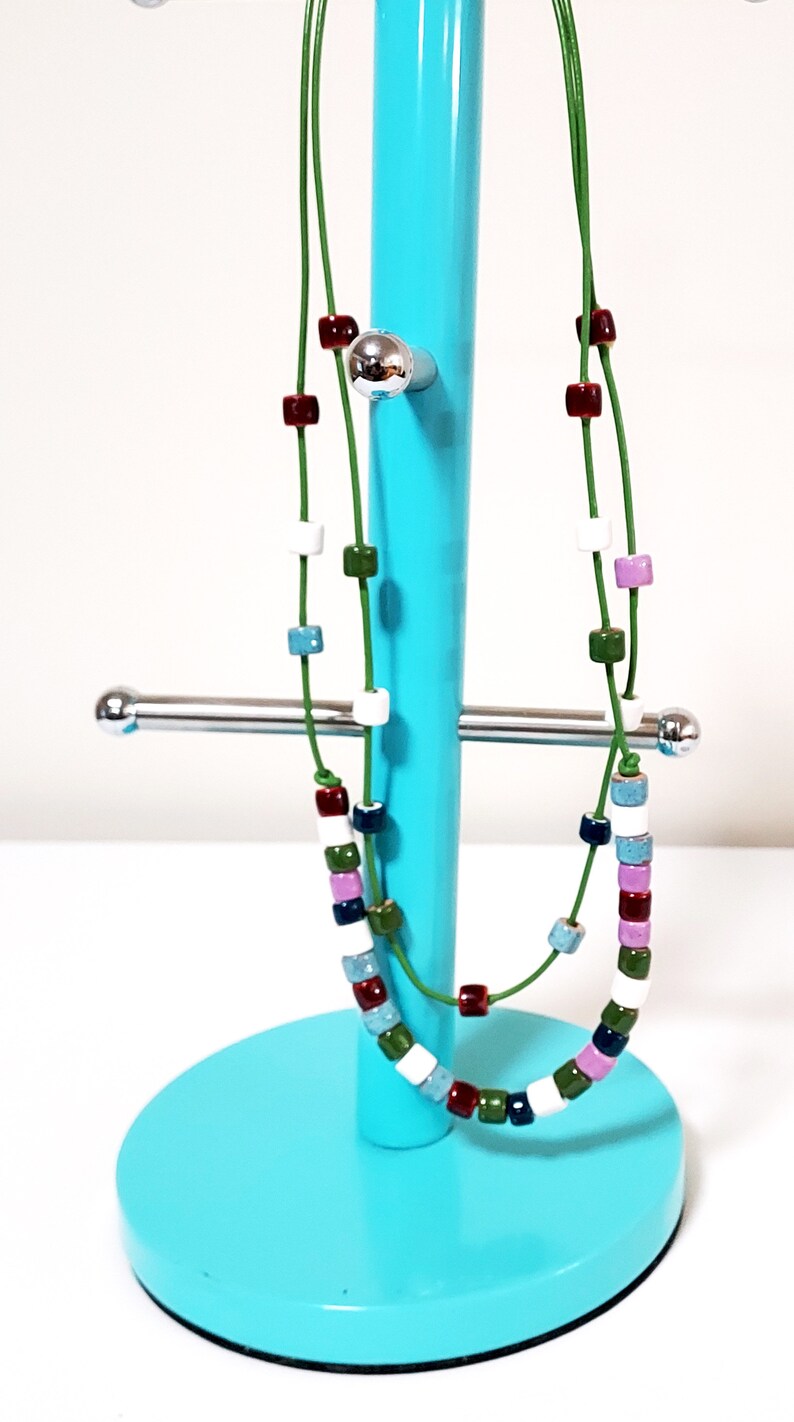 May include: A colorful necklace with square beads in various colors, including red, white, green, and blue, strung on a green cord. The necklace is displayed on a turquoise jewelry stand.