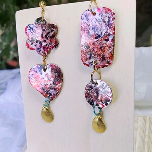 May include: A pair of unique dangle earrings. One earring features a flower and heart shape, the other a rectangle and circle. Each earring has a colorful abstract pattern in red, pink, and white, with gold-tone accents and a small seashell charm.