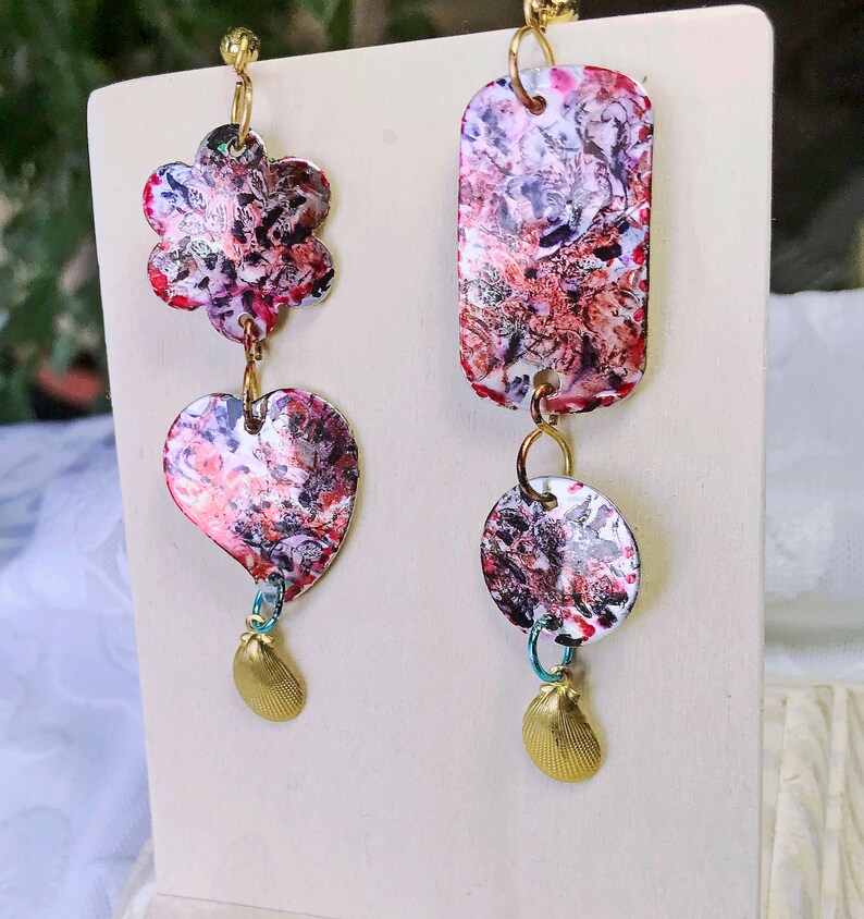 May include: A pair of unique dangle earrings. One earring features a flower and heart shape, the other a rectangle and circle. Each earring has a colorful abstract pattern in red, pink, and white, with gold-tone accents and a small seashell charm.