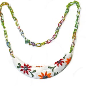 May include: A white, curved necklace pendant with red and orange floral designs and green leaves. The pendant is connected to a colorful chain with links in shades of green, white, and red. The necklace is a piece of jewelry.