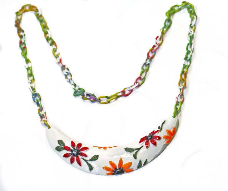 May include: A white, curved necklace pendant with red and orange floral designs and green leaves. The pendant is connected to a colorful chain with links in shades of green, white, and red. The necklace is a piece of jewelry.