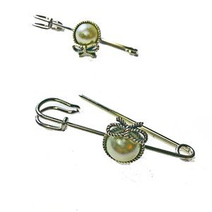 May include: Two silver-toned brooches with pearl accents. One is a straight pin with a pearl and bow detail. The other is a safety pin style with a pearl and rope-like design. Both have a classic, elegant look.