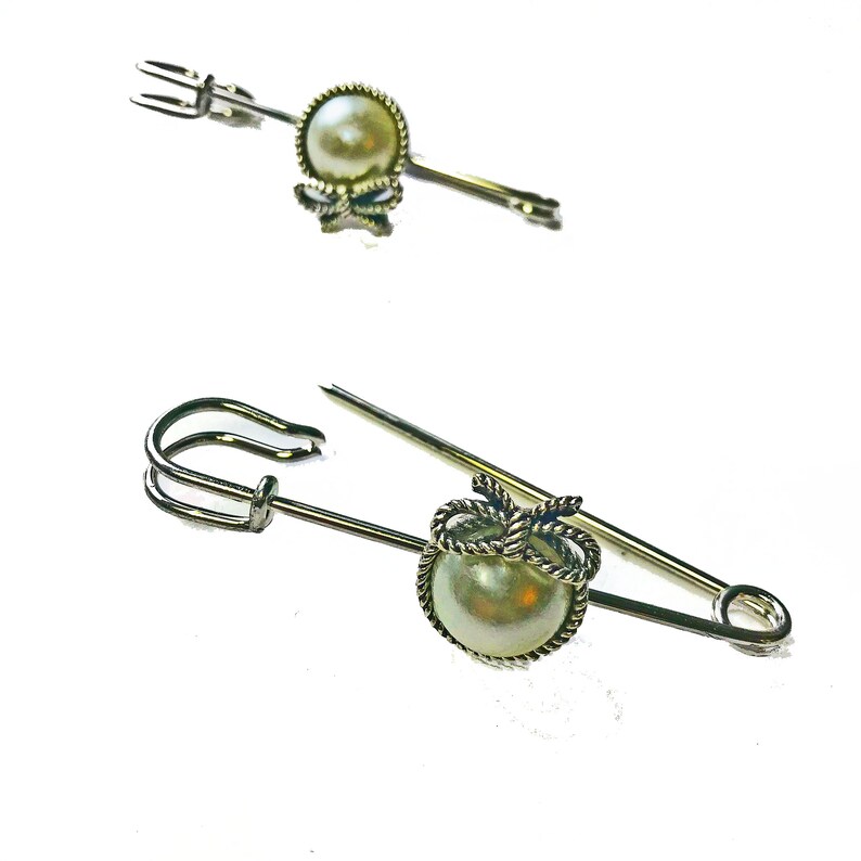 May include: Two silver-toned brooches with pearl accents. One is a straight pin with a pearl and bow detail. The other is a safety pin style with a pearl and rope-like design. Both have a classic, elegant look.