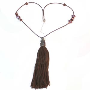 May include: A brown necklace with a long tassel pendant. The necklace features a dark brown cord with decorative beads and a silver-toned clasp. The tassel is made of a thick, textured material.