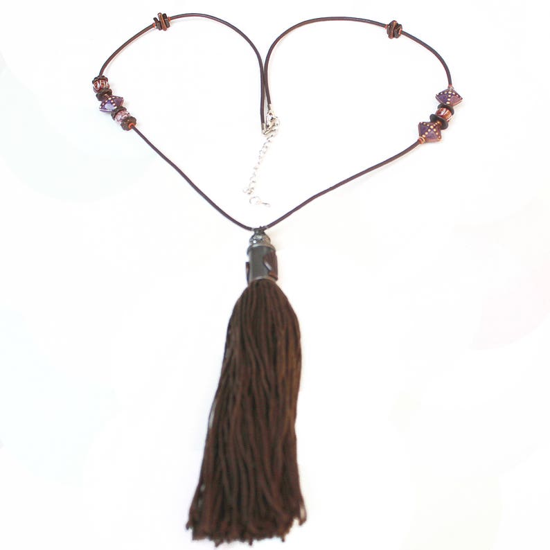 May include: A brown necklace with a long tassel pendant. The necklace features a dark brown cord with decorative beads and a silver-toned clasp. The tassel is made of a thick, textured material.