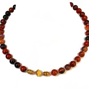 May include: A beaded necklace featuring round, reddish-brown stones. The necklace has a gold-colored centerpiece with a textured design. The beads vary in color, with some appearing darker than others. The necklace is displayed against a white background.