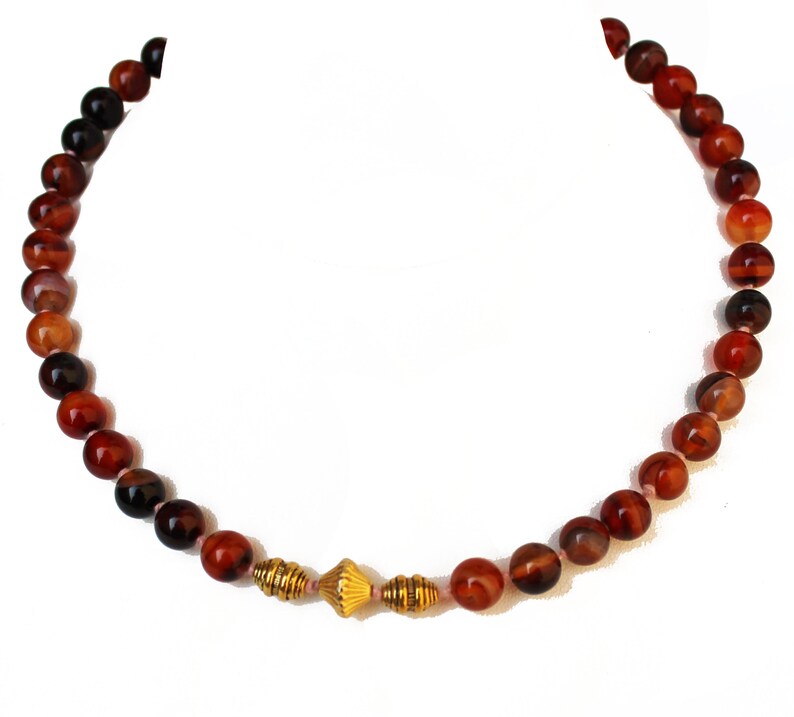 May include: A beaded necklace featuring round, reddish-brown stones. The necklace has a gold-colored centerpiece with a textured design. The beads vary in color, with some appearing darker than others. The necklace is displayed against a white background.