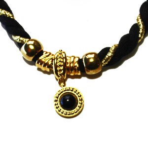 May include: A black and gold necklace with a braided cord. The necklace features gold-colored beads and a central pendant with a black stone. The pendant is surrounded by a gold-colored frame.